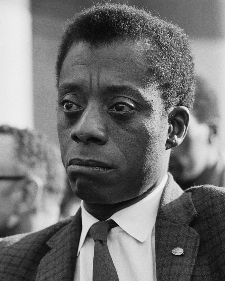Zwart-witportret van James Baldwin in pak met stropdas, peinzend opzij kijkend.